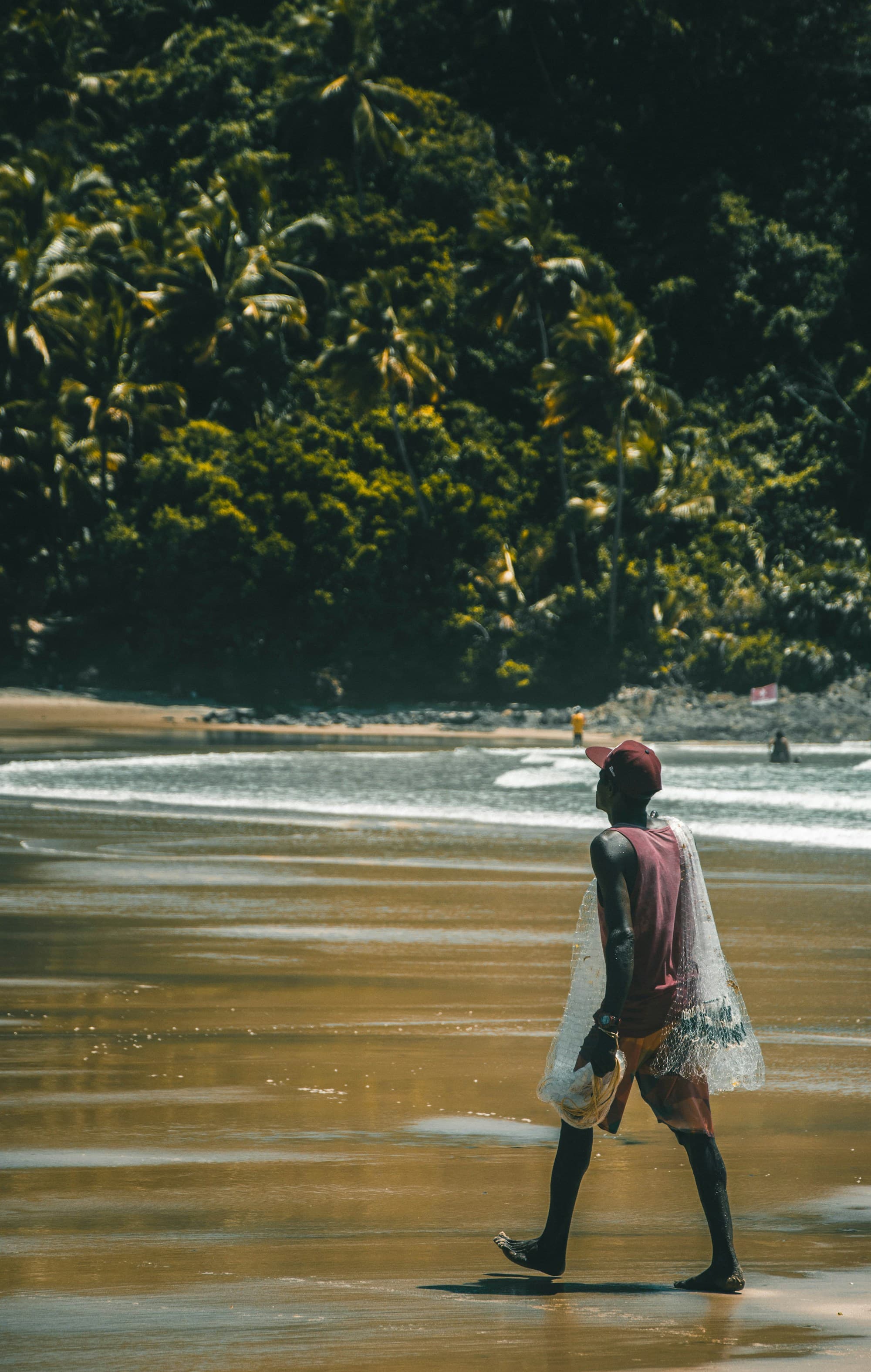 A fisher in a red cap carrying a net along the shoreline at Pescadores, Itacaré.